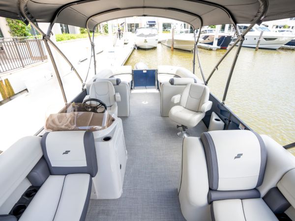 A stylish pontoon boat with cushioned seating, a center console, and a canopy, docked at a marina with calm water and other boats nearby.