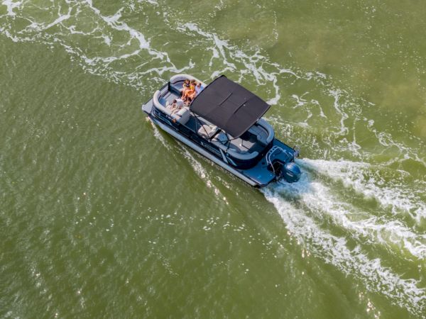 A small motorboat with a canopy cruises through greenish water, creating white wake as two people enjoy a ride.