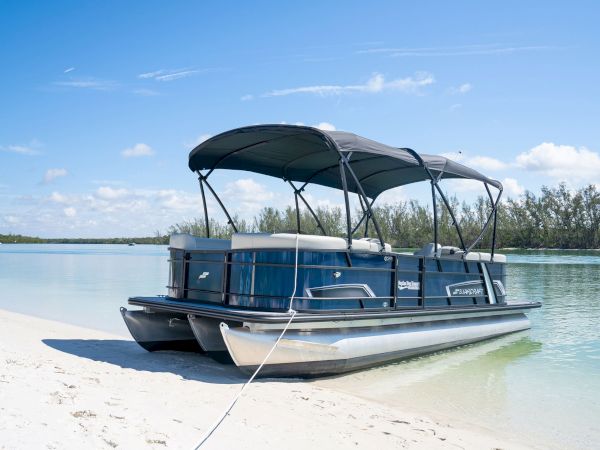 A pontoon boat docked at a sandy beach with clear blue water and a canopy, tied to shore under a bright, sunny sky.