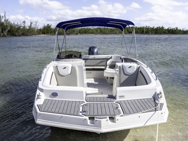 A white motorboat with gray seating sits anchored on calm water near a shoreline, under a blue canopy.