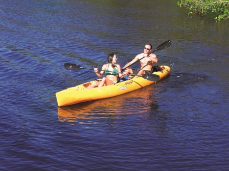 Two people in a yellow tandem kayak paddling on calm dark water near green trees, enjoying a sunny day.