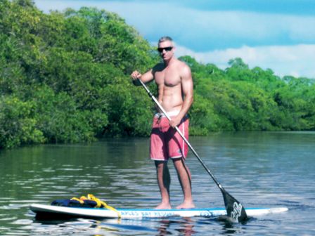 A shirtless man wearing pink shorts stands on a paddleboard, using a paddle on a calm lake with green trees in the background.