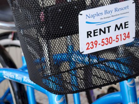 A close-up of blue bicycles with a front basket that reads &ldquo;RENT ME&rdquo; and a phone number, advertising bike rentals at Naples Bay Resort.