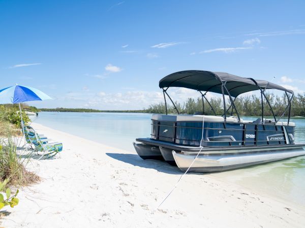 A sunny beach with white sand, calm turquoise water, a blue boat with a canopy anchored near shore, and a blue umbrella on the beach.