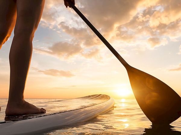 A person stands on a paddleboard at sunset, holding a paddle over calm water. The horizon glows with warm orange light.