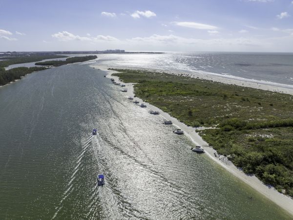 Aerial view of a river/lake mouth with a narrow channel, boats cruising, and a sandy coast with green vegetation along the shoreline.