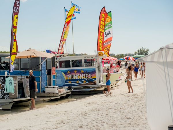 Beach scene with colorful flags on trailers and tents, people walking along sandy shore near a row of vans and vendors.