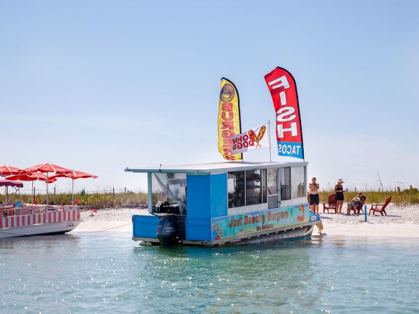 A blue docked boat on a calm, shallow beach with red umbrellas, flags, and people relaxing by the water&rsquo;s edge, ready to serve refreshments.