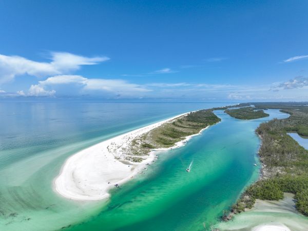 A turquoise coastal scene with white sandy bars forming a curved spit, shallow lagoons, and clear blue skies over calm tropical water.