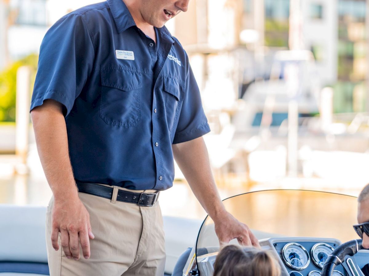 A man in a navy shirt greets a child on a boat, with a dashboard and another person seated nearby in a sunny marina setting.