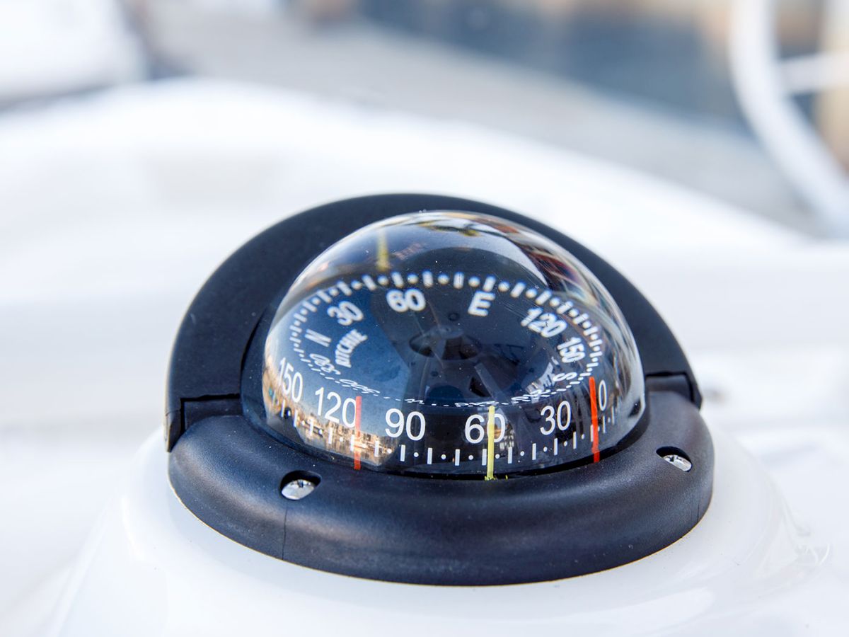 A close-up of a boat compass showing degrees, with a black mounting base, on a white surface near clear water and railing.