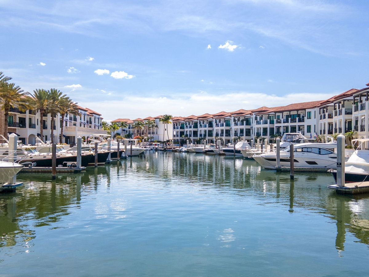A sunny marina lined with boats and white buildings, calm water reflecting the blue sky, palm trees, and a peaceful vacation vibe.