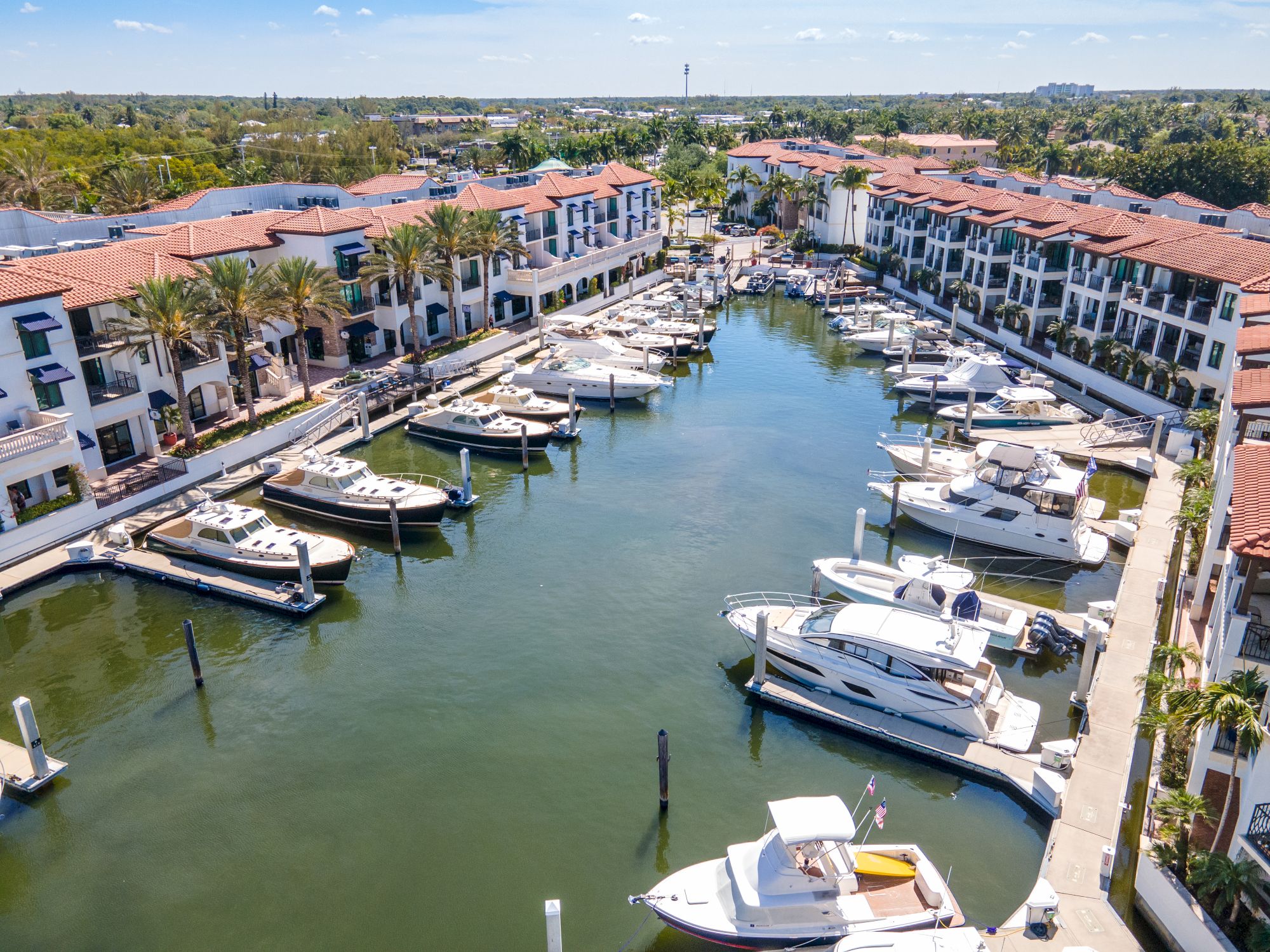 Aerial view of a marina in a sunny coastal complex, with boats docked along a canal lined by palm trees and red-roofed buildings, tranquil blue water.
