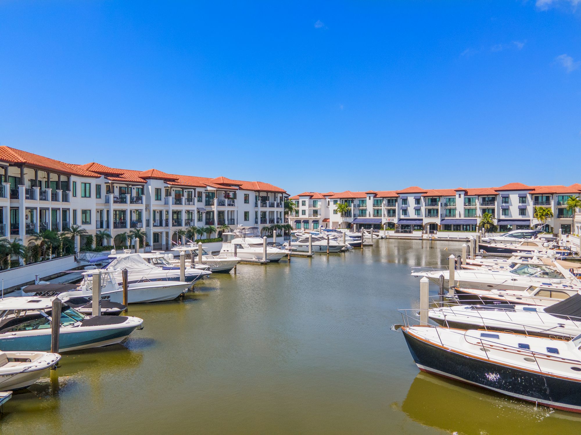 A marina surrounded by white buildings with red-tiled roofs, boats docked along calm water under a clear blue sky.