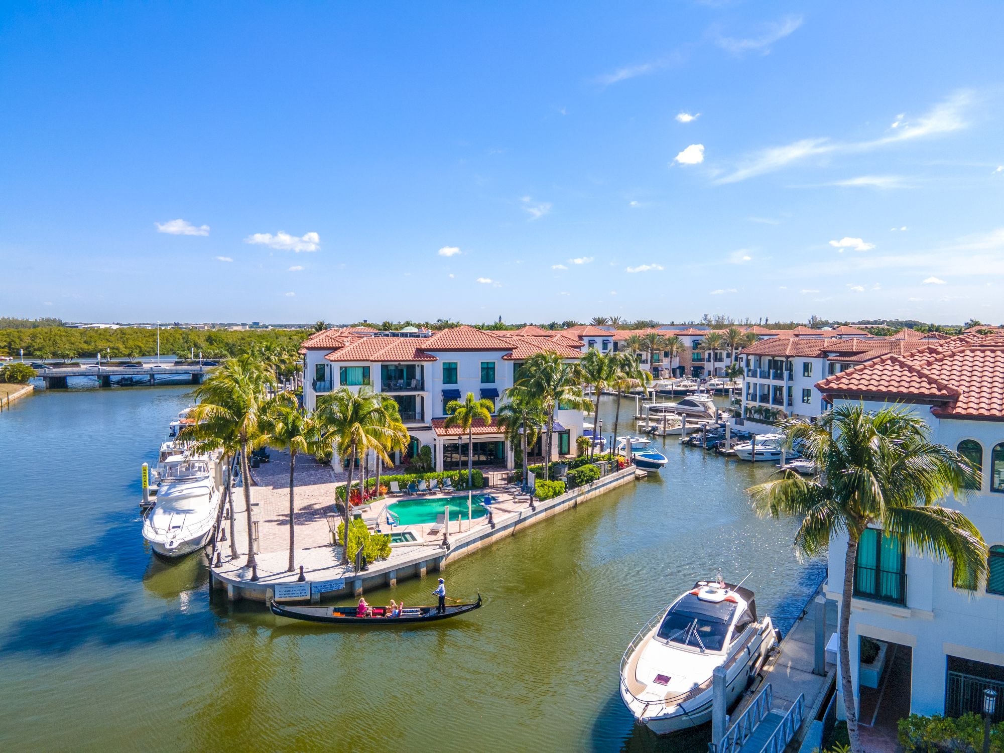 A sunny waterfront with canal homes, boats docked along slips, palm trees, and a bright blue sky over a marina community.