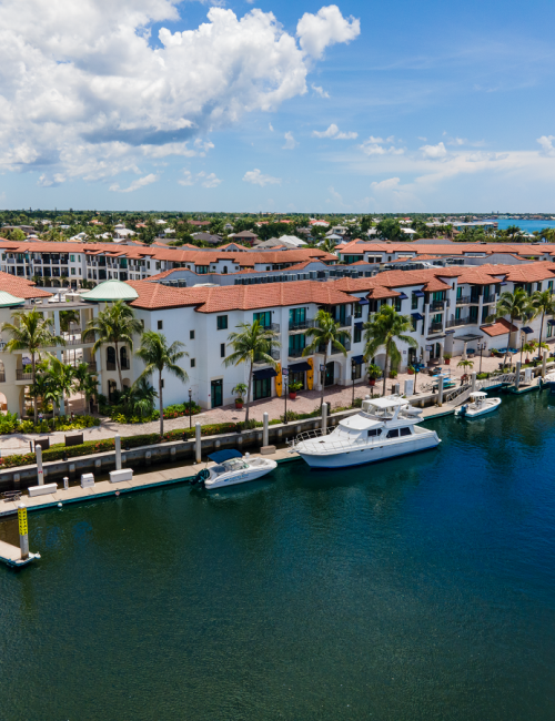 Colorful waterfront townhouses line a canal, with palm trees, boats docked along a calm marina under a bright blue sky.