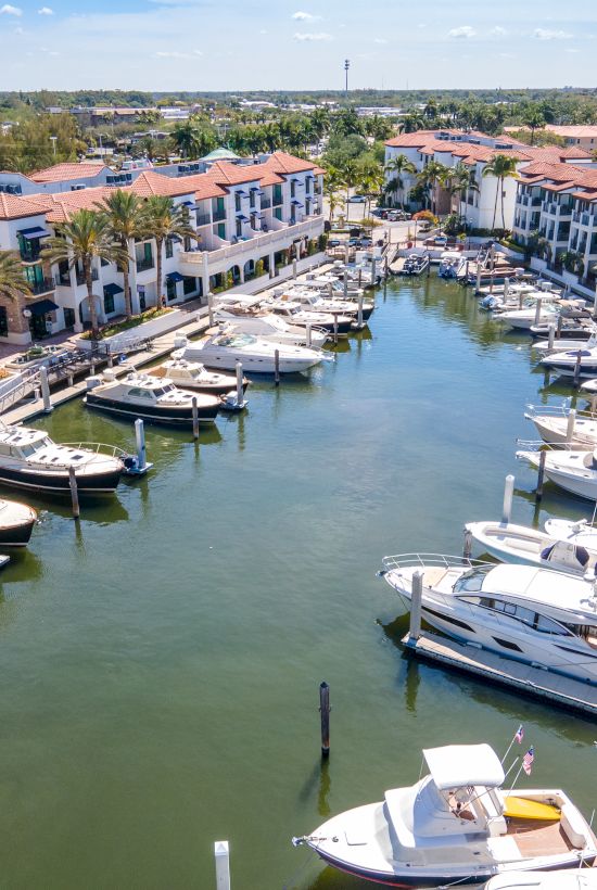 A canal lined with luxury townhomes and private docks, filled with yachts and boats, palm trees, and calm water in a sunny coastal setting.