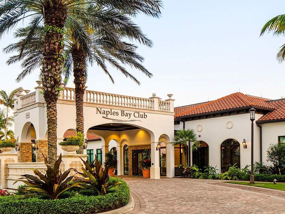 A sunny resort entrance with palm trees and a Mediterranean-style building labeled &ldquo;Naples Bay Club,&rdquo; inviting drive and lush landscaping.