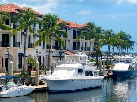 A marina with palm trees, boats, and a waterfront building complex by calm water.
