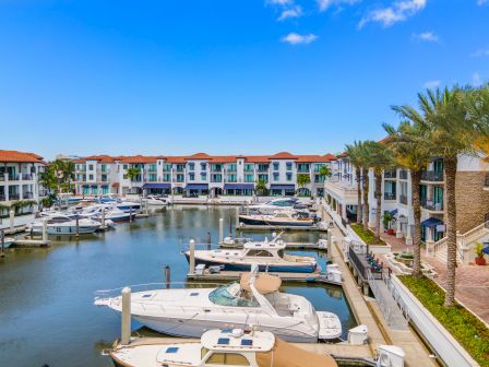 A marina with several white boats docked along calm water, surrounded by colorful low-rise buildings and palm trees under a blue sky.