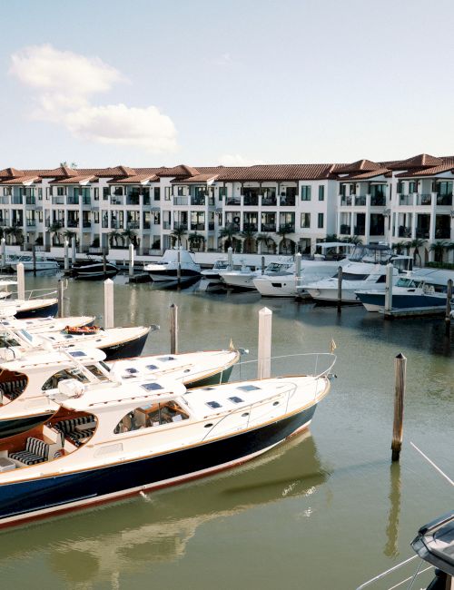 A marina with numerous docked boats and a row of white, multi-story townhouses along the water, under a bright, partly cloudy sky.