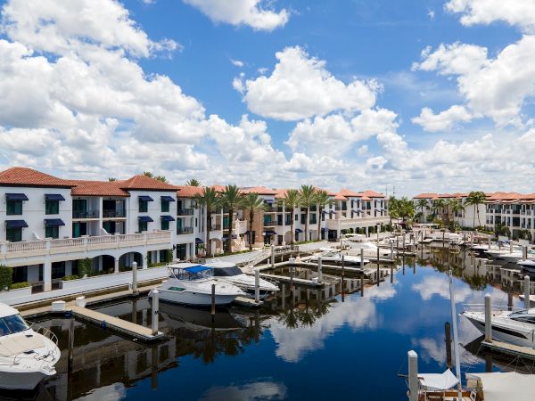 A sunny marina with calm water reflecting buildings, boats, and palm trees lining a row of beige and white apartment-style docks.