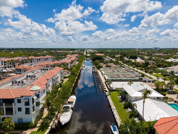 Aerial view of a canal lined with boats, tropical buildings on both sides, palm trees, clear blue sky with fluffy clouds, and waterfront homes.