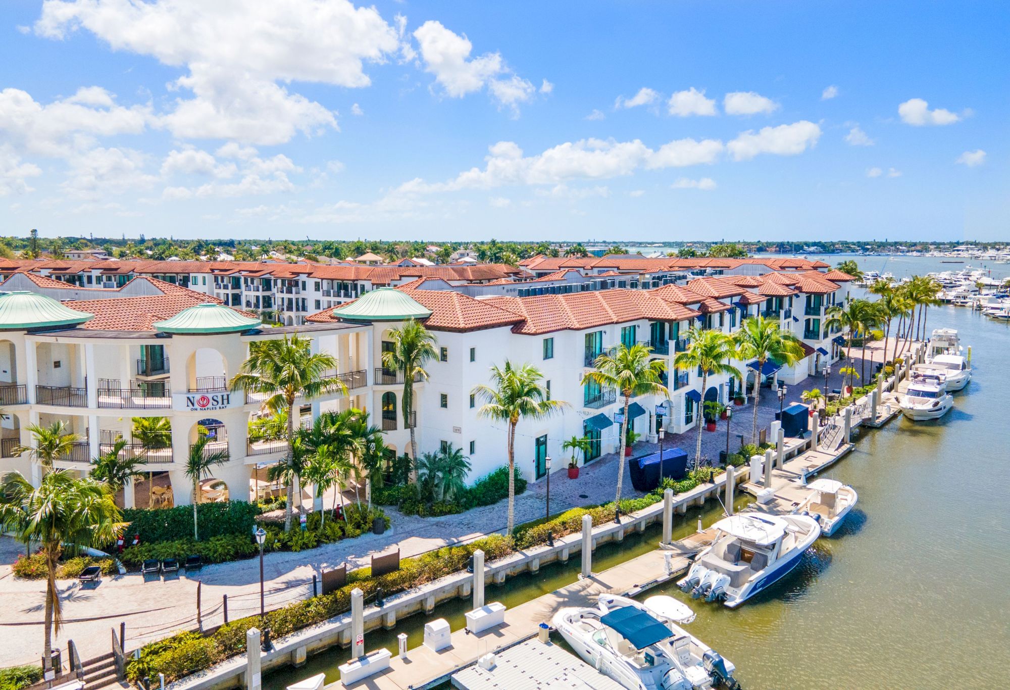 Colorful Mediterranean-style townhouses along a sunny canal with palm trees and boats docked at the waterfront, a bright, inviting scene.