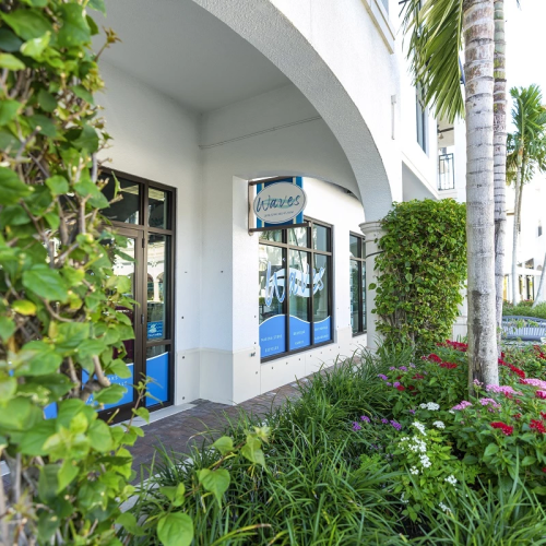 A sunny resort courtyard with arched entryway, palm trees, and colorful flowerbeds along the walkway.