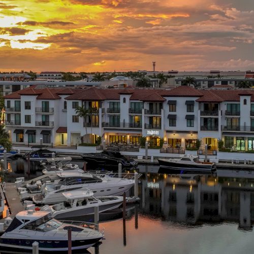 Sunset over a marina with yachts and a white resort building, glowing clouds reflecting on calm water.