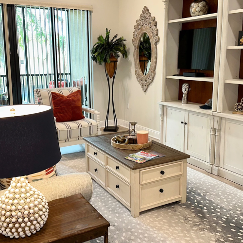 A bright living room with a sofa, coffee table, window views, and a white TV/fireplace console decorated with plants and vases.