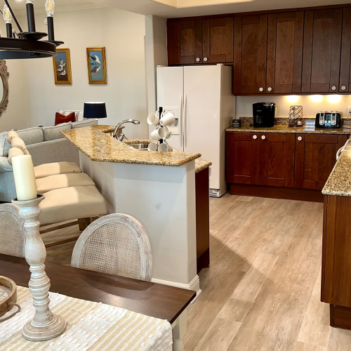 A modern living room and kitchen with granite island, dark wood cabinets, coffee makers, and a sofa in warm lighting.