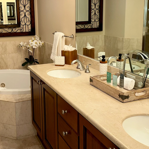 A bright hotel bathroom with a vanity sink, toiletries on the counter, and a bathtub and towels in view.