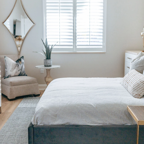 A bright modern bedroom with a bed, nightstand, armchair, dresser, potted plant, and diamond mirror by sunny windows.