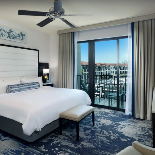 A modern hotel bedroom with a king bed, TV, and a balcony view through glass doors.