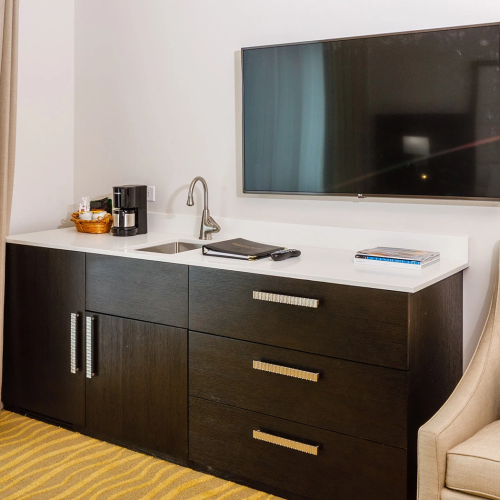 Hotel room vanity with sink and coffee maker, large flat-screen TV on wall, beige chair and striped carpet.