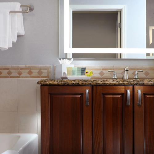 A modern bathroom vanity with a granite countertop, sink faucets, folded towels, a window, and a leafy vase on the counter.