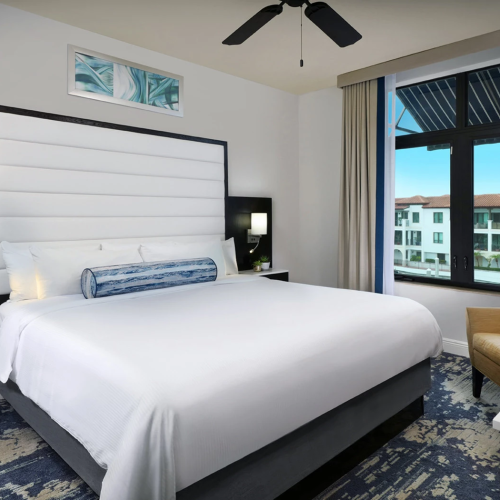 A modern hotel bedroom with a large bed, bedside lamps, and a view through big windows, with a ceiling fan overhead.