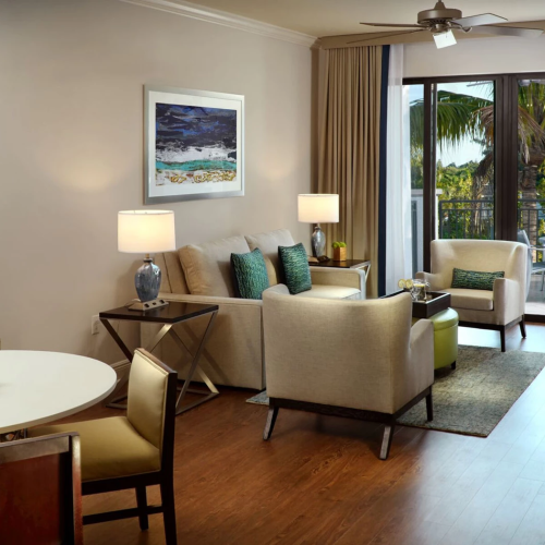 A bright living room with beige sofas, lamps, curtains, a round table with fruit, and sliding doors to a tropical view.