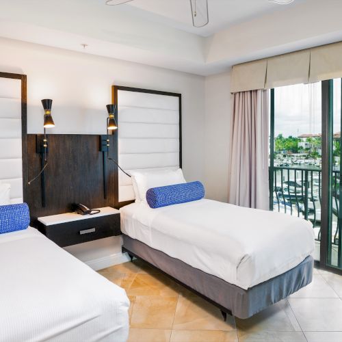 A modern hotel bedroom with two twin beds, bedside lights, and beige curtains over balcony doors.