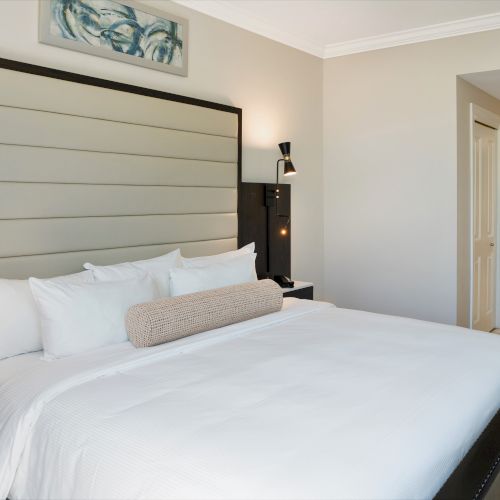 A modern bedroom with a neatly made bed, bedside lamps, wall art, and a doorway leading to a tiled bathroom.