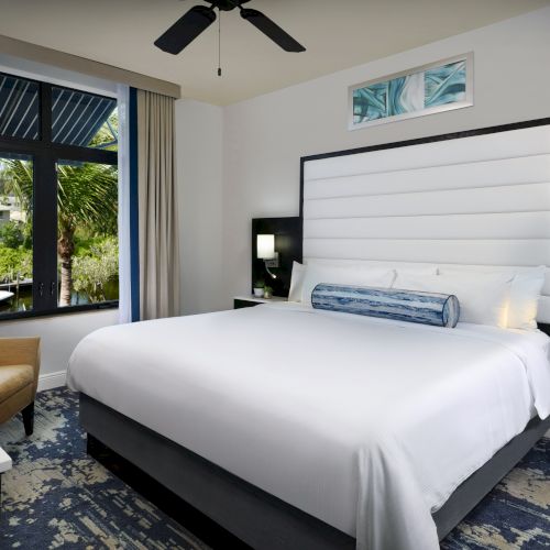 A modern hotel bedroom with a large bed, bedside lamps, a ceiling fan, curtains, and a view through big windows.