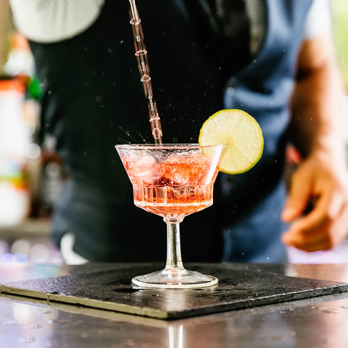 A bartender pours a pink cocktail into a glass with ice, while lime slices fly through the air.