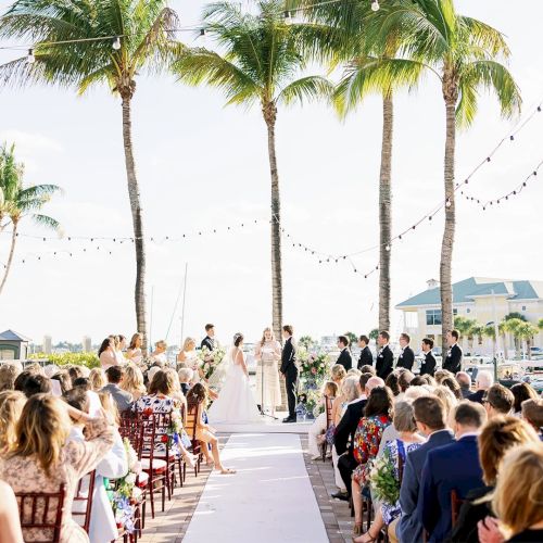 An outdoor wedding ceremony on a palm-lined deck with the bridal party up front, surrounded by cheering guests.