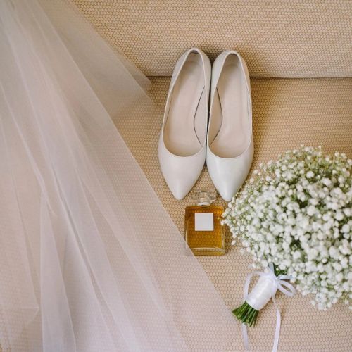 A wedding detail shot: white bridal heels, a bottle of perfume, and a white-green bouquet on a textured couch with draped fabric.
