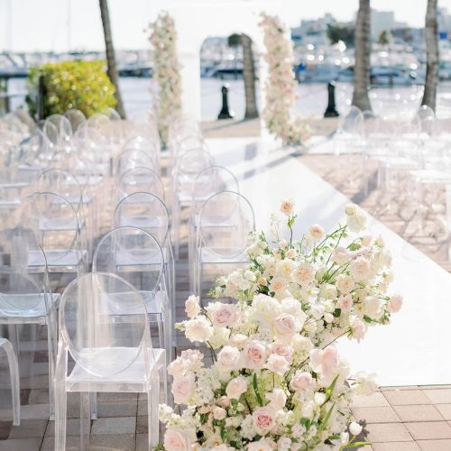 An outdoor terrace wedding setup with clear chairs, a white-draped aisle, and a bouquet of pastel flowers by the railing.