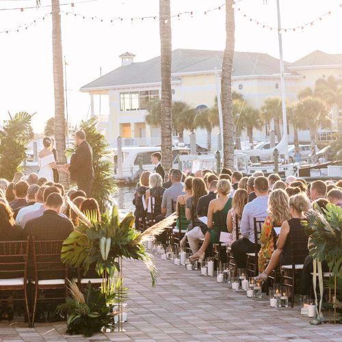 An outdoor wedding ceremony on a terrace with rows of guests, palm trees, and lit candles at sunset.