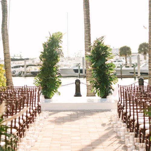 A waterfront wedding aisle with red chairs, glowing palm trees, and boats in the marina behind, set for a ceremony.