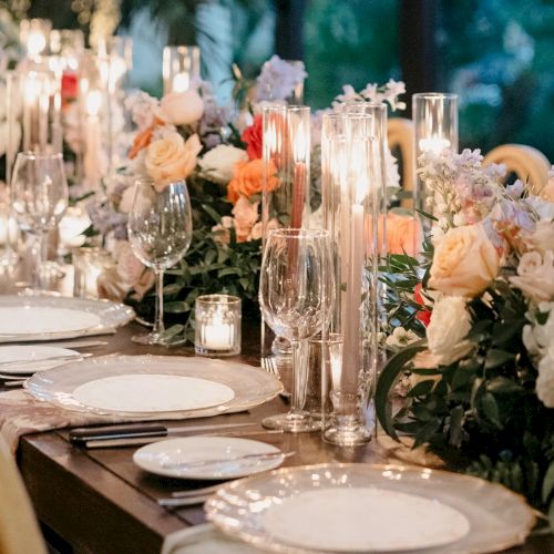 A reception table set for a celebration, with candles, glassware, and colorful floral centerpieces.