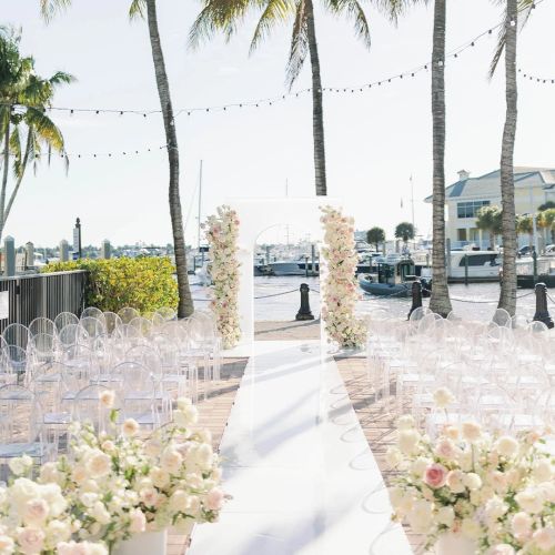 Outdoor waterfront wedding setup with white chairs, floral arch, palm trees, and boats in the background, under string lights.